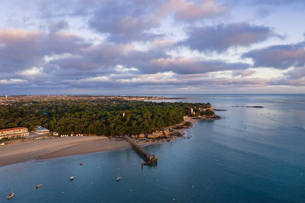 Sakin bir kıyı şeridi manzarası, mavi suya uzanan ahşap iskele, kumlu sahil, ve yumuşak günbatımı bulutları altında kıyı boyunca verimli çam ormanı..