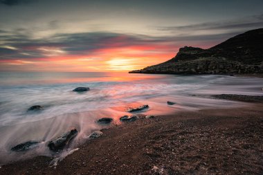 Sersemletici gün doğumu dalgalar kayaları ve karanlık çakıl taşlarını yıkarken ufku boyar. Sıcak tonlu sakin bir sahil manzarası, uçurum silueti ve yansıtıcı su, Costa Almeria, İspanya