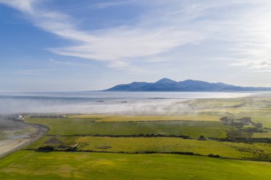 Sakin bir kıyı manzarası canlı yeşil alanlar, sisli bir kıyı şeridi ve uzak mavi dağları gösterir. Berrak gökyüzü ve sakin su, doğa, seyahat ve açık hava yaşam tarzları için mükemmel bir manzara yaratır..