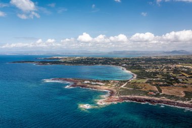 Güneşli bir sahilin geniş hava manzarası: berrak turkuaz su, engebeli kıyı şeridi, küçük beyaz binalar, Sardunya, İtalya