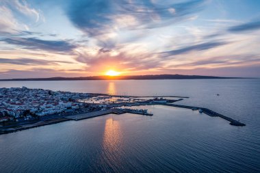 Alacakaranlıkta sakin bir liman kıyı kenarı, rıhtımlar boyunca sıralanmış tekneler ve ufukta batan güneş sakin sulara sıcak bir yansıma yapıyor.