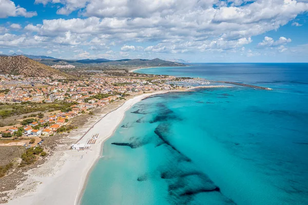 Drone view of a coastal town along a turquoise sea, white sand beach, colorful houses, and a long pier, Sardinia, Italy