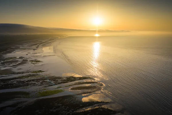 A tranquil seascape at sunset features a glowing horizon, gentle waves, and reflective tidal pools along the shore. Warm light bathes the water and beach, evoking peace, serenity, and natural beauty, Newcastle, Northern Ireland