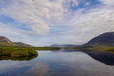 Huzurlu bir dağ gölü, tüylü bulutları ve mavi gökyüzünü yansıtır. Orman kıyıları ve uzak tepelerle çevrili, Galway, İrlanda