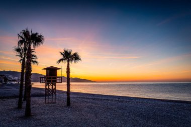 Bir çakıl taşı plajında sakin bir günbatımı, sakin bir denize bakan palmiye ağaçları ve bir cankurtaran kulesi, İspanya Castell de Ferro,