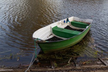 Küçük yeşil bir kayık gölün kenarında dinleniyor, halat ve zincirle bağlanmış, sakin dalgalı su ve doğal bitki örtüsüyle çevrili..