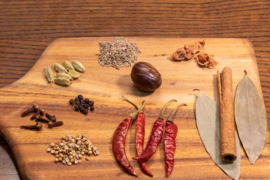 This image showcases a beautifully arranged selection of spices and herbs on a wooden cutting board. The assortment includes chili peppers, cinnamon sticks, bay leaves, cardamom pods, cloves, nutmeg, ginger, and various seeds and grains.