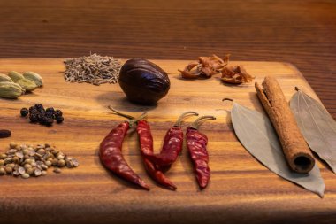 Bu resimde, acı biber, tarçın çubukları ve kakule kapsülleri de dahil olmak üzere ahşap bir kesme tahtasının üzerinde çeşitli baharat ve otlar sergilenmektedir. Malzemelerin canlı renkleri ve dokuları görsel olarak çekici bir kompozisyon oluşturuyor..