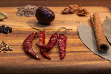 Bu resimde, kırmızı biber, tarçın çubukları ve çeşitli otlar da dahil olmak üzere çeşitli baharat ve malzeme içeren ahşap bir kesim tahtası yer alıyor..