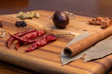 Bu resim, acı biber, tarçın çubukları, kakule ve defne yaprağı da dahil olmak üzere çeşitli baharat ve malzemelerle dolu ahşap bir kesim tahtasını tasvir ediyor. Kesme tahtası bir masaya konur ve arka plan bulanıklaşır..