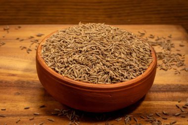 Bu resim ahşap bir yüzeye yerleştirilmiş kimyon tohumlarıyla dolu küçük bir terracotta kasesini gösteriyor. Kase çerçevenin ortasında ve kimyon tohumları masaya hafifçe akıyor. Ahşap yüzeyin doğal bir tahıl deseni var..