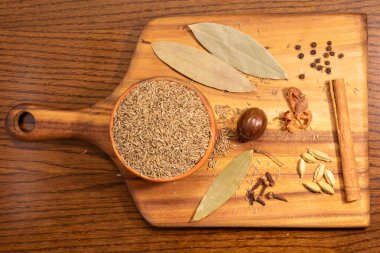 Bu resim, ahşap bir masanın üzerinde bir kase pirinç de dahil olmak üzere çeşitli baharat ve otlarla dolu ahşap bir kesim tahtasına sahip. Kesme tahtası açık renkli ahşaptan yapılmış ve sapında bir delik var..