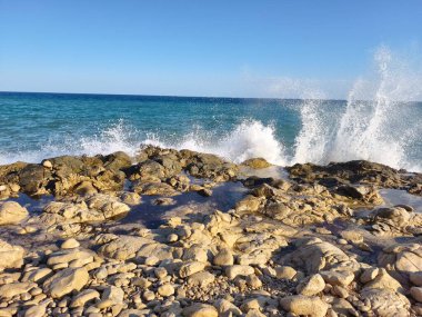 Experience the stunning view of ocean waves crashing against the rocky shore under a clear blue sky