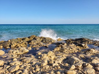 Waves crash against rugged rocks under vast blue skies, creating a breathtaking coastal scene of beauty