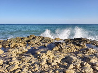 Witness the stunning view of waves crashing gracefully against the rocks on a clear day by the beautiful sea