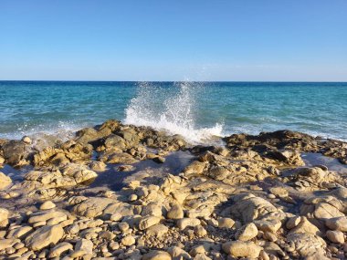 The beautiful ocean waves are crashing powerfully against the rocky coastline beneath a stunning clear blue sky