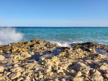 A tranquil seascape captures the moment as waves crash powerfully against a rugged coastline beneath a clear blue sky