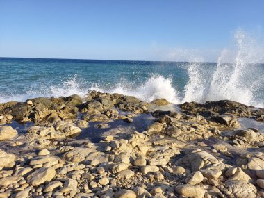 Experience the stunning view of ocean waves crashing against the rocky shore under a clear blue sky
