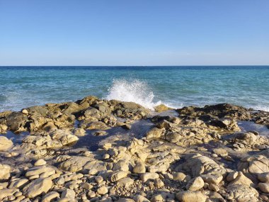 Waves crash against rugged rocks under vast blue skies, creating a breathtaking coastal scene of beauty