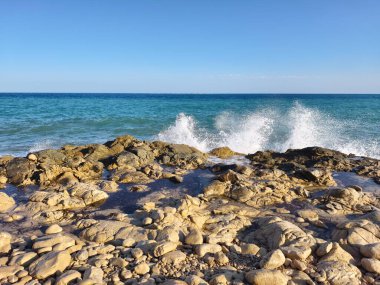 Witness the stunning view of waves crashing gracefully against the rocks on a clear day by the beautiful sea