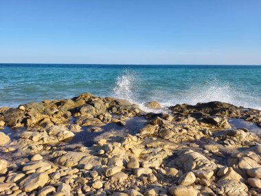 A tranquil beach view with shimmering waves lapping against rocky shorelines under a clear blue sky