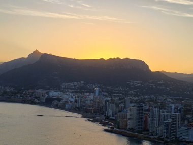 Günbatımı şehir silüeti, şehir kıyı şeridi üzerinde altın gün batımı, dağ ve okyanus manzaralı sakin bir akşam.