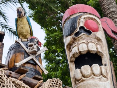 Palmiye yapraklı, tropik festival manzarasının ortasındaki yıpranmış korsan sembollerinin üzerindeki renkli papağan.