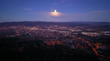 Şehir manzarası aydınlık ayın altında ışıldıyor, Parlayan mahalleler ve uzak tepelerle dolu canlı bir gece sahnesi