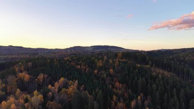 Geniş açılı panoramik görüntüler, sakin günbatımını yumuşak pastel tonlarıyla tepelik orman manzarasının üzerine yansıtıyor.