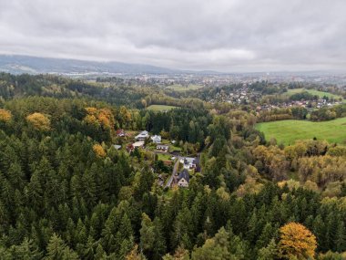 Yuvarlanan tepelerin yakınındaki ormanda gizlenmiş bir hava köyü. Karışık tepe örtüsü ve sonbahar tonlarının çerçevelediği evler kümesi.,