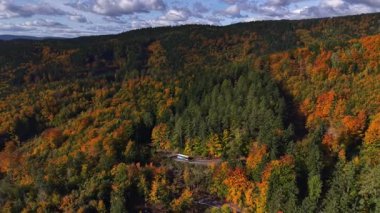 Sakin kırsal patikalar canlı yapraklı ağaçlar ve uzak dağ manzaraları arasında zikzak çiziyor.