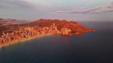 Hava şehri kıyıları gün doğumunda, ılık ışık banyoları yüksek binalar ve dağlı kumlu plajlar.