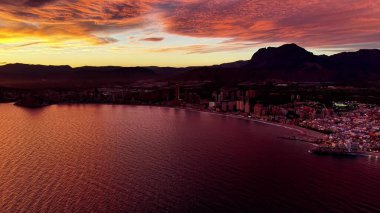 Kıyı kenti üzerinde alacakaranlık, panoramik hava aydınlanan sokaklar, liman kenarı ve dağ silueti