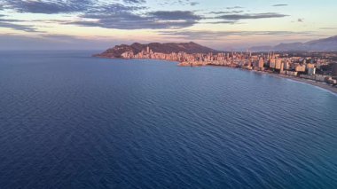 Kayalıklarda ve okyanuslarda sıcak güneş ışığı, yumuşak pastel gökyüzü, uzak kentsel şeritler...