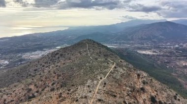 Hava sırtı yaklaşımı uzak kıyı şeridini ve vadiyi gösteriyor, virajlı toprak yolları kayalık çalıların arasından geçiyor.,