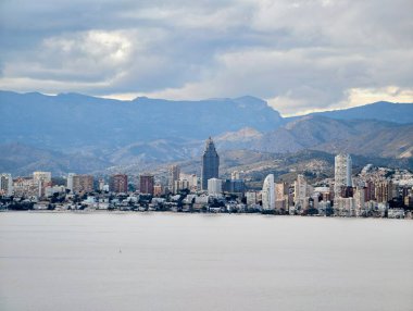 Ayırt edici kulesi ve dağ zeminiyle gökyüzü manzaralı geniş sakin bir deniz önplanda ve katmanlı