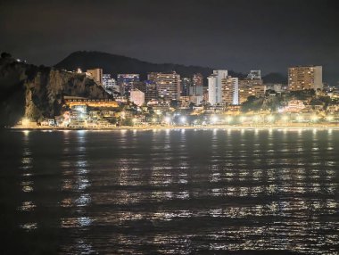 Gece kıyı şeridi yansımaları, ışıltılı rıhtım ışıkları ve dağ silueti ile sakin