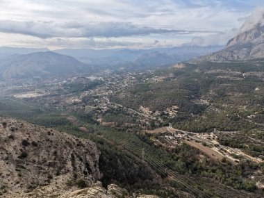 Hava bulutlu tepe görüntüsü insansız hava aracının çektiği sarmal izler, dağınık evler ve uzak sahil manzarası.