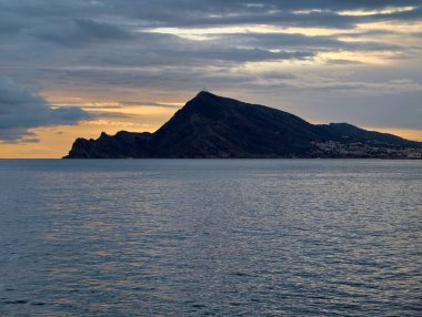 Karanlık adalı karamsar deniz manzarası, dalgalı suyun üzerinde ağır bulutlar, uzak kıyı ışıkları