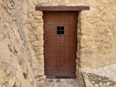 Taş duvarda çivilenmiş ahşap kapı, ağır demir çiviler ve küçük kare gözetleme deliği, desenli duvar ustalığı.