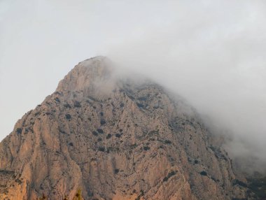 Alçak bulutların altında gizlenmiş granit zirve pürüzlü dokuyu ve ılık tonları gözler önüne seriyor, yakın kaya yüzeyi