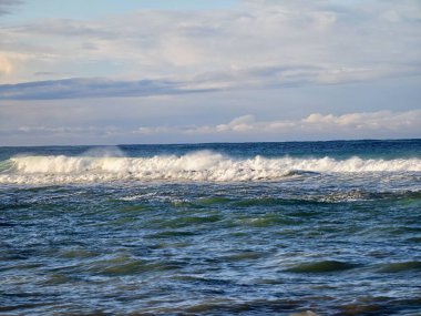 Mavi okyanus dalgaları ufka doğru yuvarlanıyor, yumuşak bulutlu gökyüzü altında köpüklü dalgalar, yumuşak dalgalar şekilleniyor.