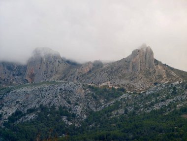 Alçak bulutlar ve sislerin arasından yükselen ikiz engebeli tepeler, çam kozalaklı yamaçların çerçevelediği kireçtaşı sırtlarını süpürüyor.