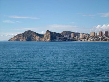 Kıyı binaları ve turkuaz denizi, dar kıyı şeridi ve parlak gökyüzü altında hafif dalgalar,