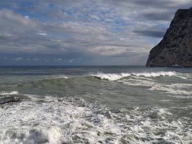 Burun dalgaları kayalık kayalıklara vuruyor ve köpüklü sörfler, engebeli kıyı şeridi profili ve bulutlu altında dik taş yüzler.