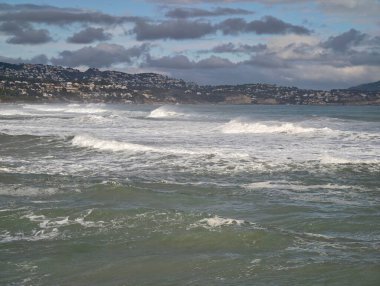 Körfez manzaralı dalgalar ve kıyı şeridindeki uzak kasaba, limanın yukarısında yumuşak bulutlar ve soluk renkler yaratıyor