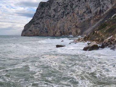 Çalkantılı denizin yanında kayalık, fırtına bulutları geliyor, köpüklü köpükle kayalara çarpıyor.