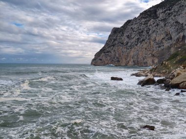 Kaynamış sörfün ön görüntüsü, kayalık kıyılara doğru akan köpüklü su, bulutlu gökyüzü ve dinamik tepeler oluşuyor.