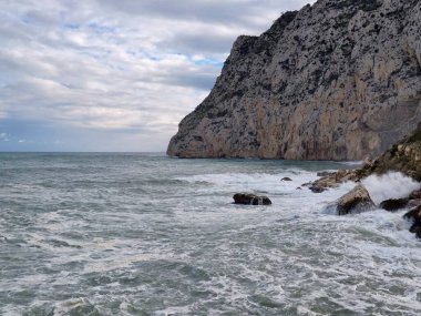 Dik kireçtaşı kayalıklar sörfe bakıyor, köpüklü dalgalar kırbaçlanıyor, görünür katman ve yıpranmış dokular, dramatik.