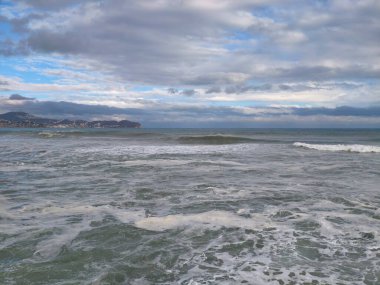 Geniş kıyı manzarası, uzak burunlu, dalgalı deniz ve katmanlı bulut örtülü, sakin ama dramatik